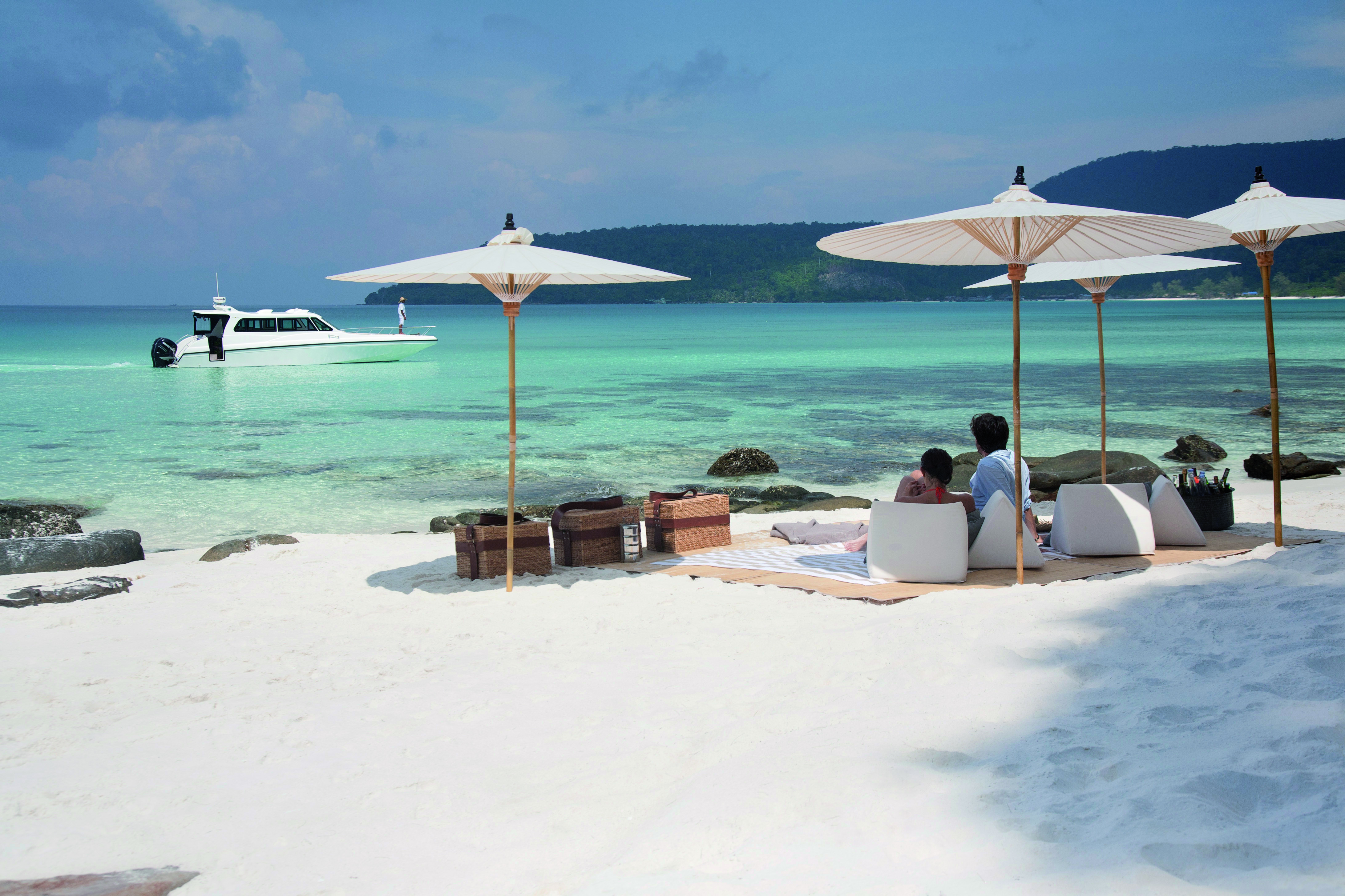Picnic of the neighbouring island of Koh Rong with white sandy beaches leading onto a clear turqouise sea with white umbrellas and wooden picnic baskets on the sand