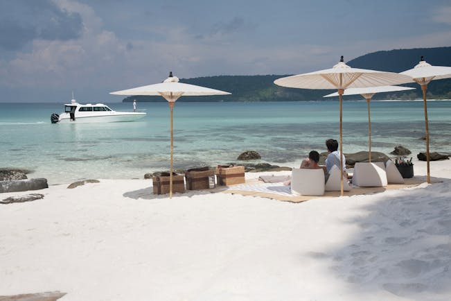 Picnic of the neighbouring island of Koh Rong with white sandy beaches leading onto a clear turqouise sea with white umbrellas and wooden picnic baskets on the sand