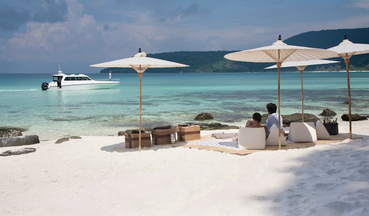 Picnic of the neighbouring island of Koh Rong with white sandy beaches leading onto a clear turqouise sea with white umbrellas and wooden picnic baskets on the sand