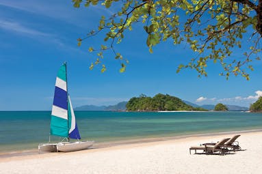 Andaman Langkawi white sandy beach with loungers and sailboat with blue and white sail, islands in distance