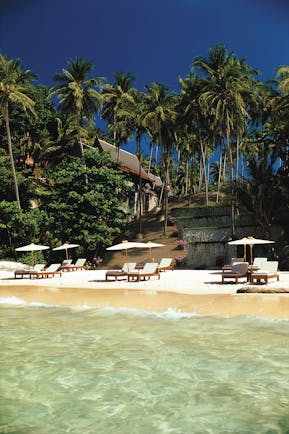 Beach with clear blue sea, sun loungers and umbrellas on the sand and large palm trees in the sea