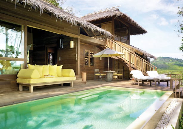 Exterior view of a deluxe villa at the Six Senses Yao Nai with a large rectangular pool with wooden decking area, seating areas with beach hut style buildings to the side