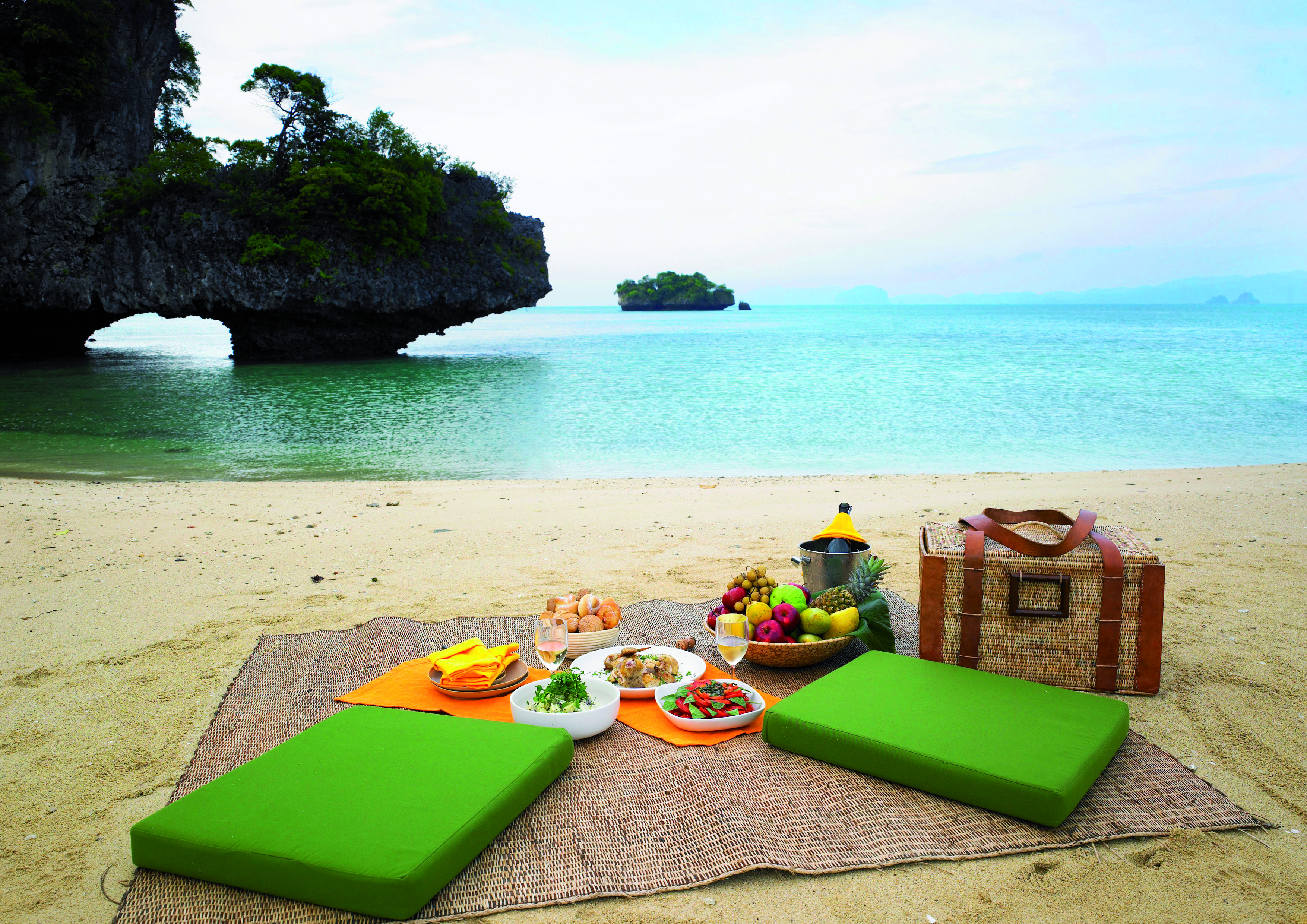 Picnic laid out on sandy beach with green seating cushions, a picnic basket and blue sea in the background