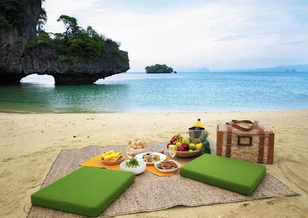 Picnic laid out on sandy beach with green seating cushions, a picnic basket and blue sea in the background