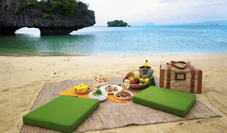 Picnic laid out on sandy beach with green seating cushions, a picnic basket and blue sea in the background