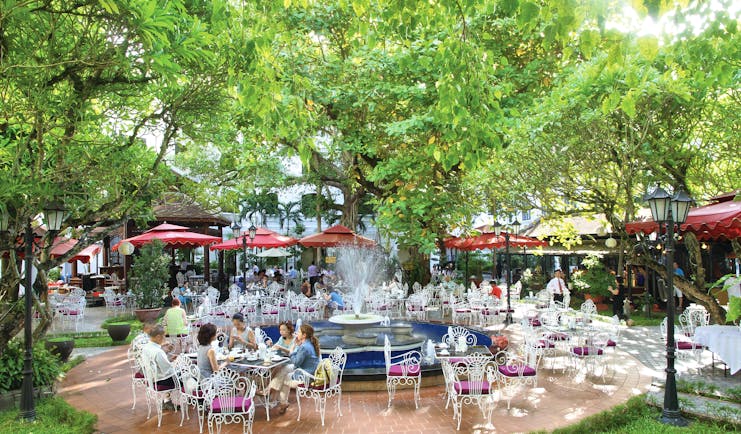Restaurant at the Saigon Morin Hotel in Vietnam with outdoor seating areas shown beneath thick green trees, white chairs and red umbrellas 