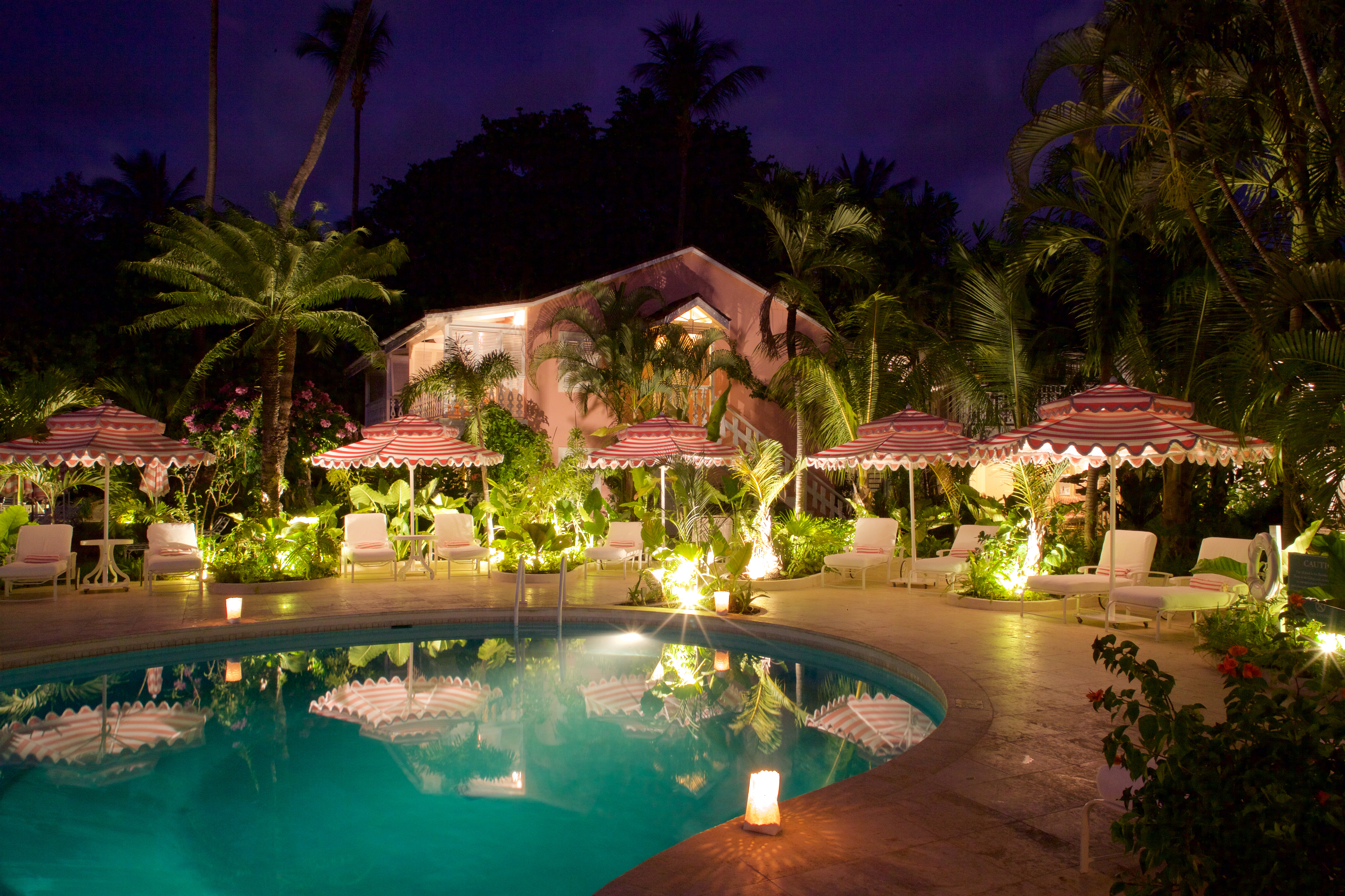 Pool at night with pink hotel exterior building in background, pink and white umbrellas and pool lights 