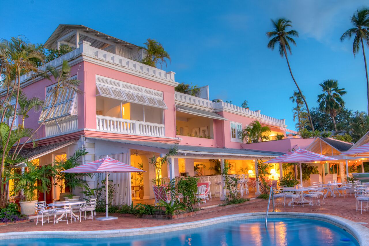 Outdoor swimming pool with pink umbrellas and deck chairs set up and pink hotel building in backgroun