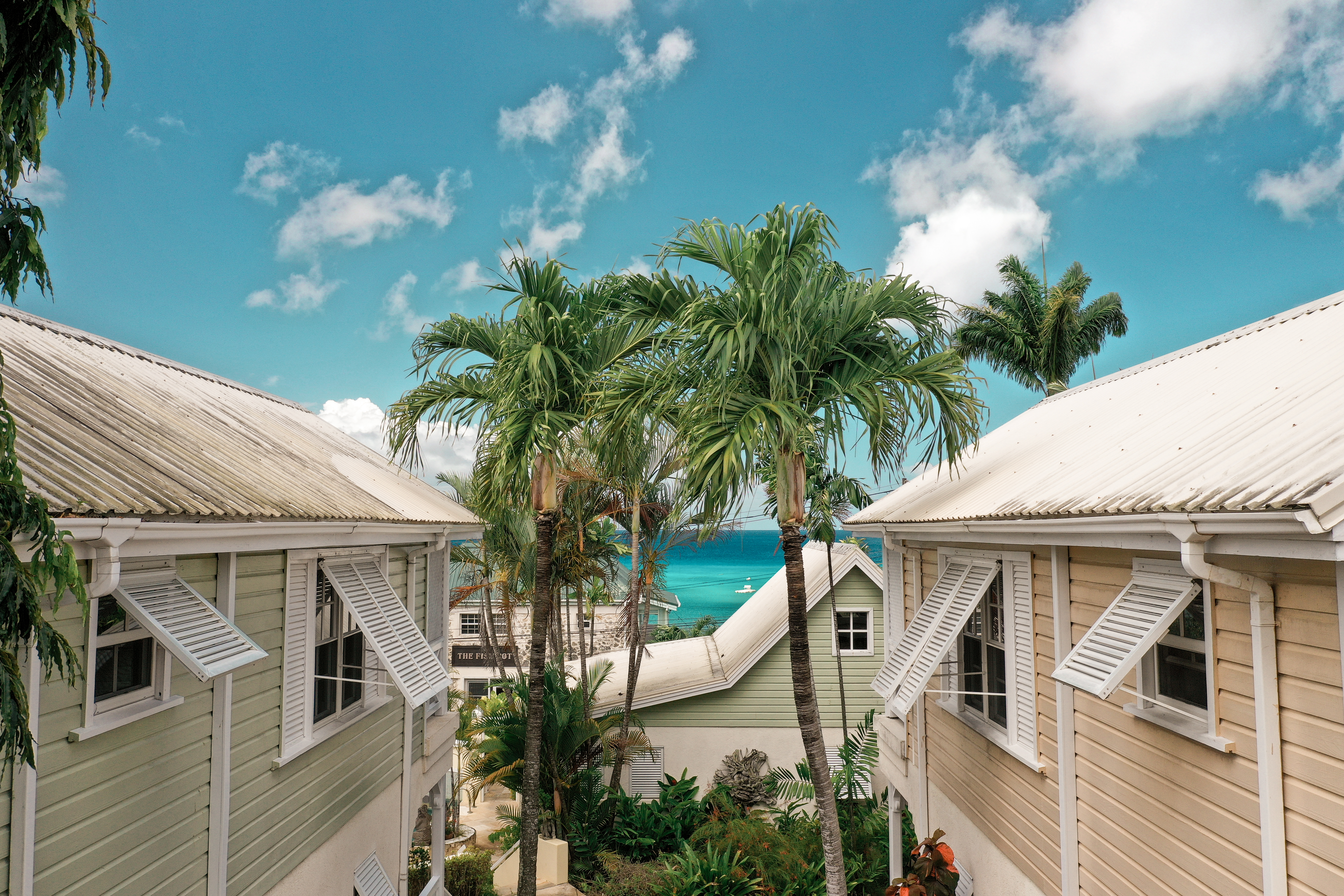 Little Good Harbour seen from an elevated angle, with woodn-clad buildings, shuttered windows, wooden roofs, and palm trees, with the sea visible in the distance