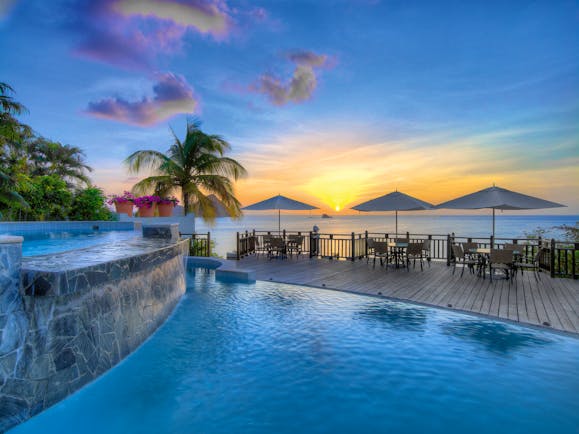 View of the pool at sunset with palm trees and chairs around and yellow sun setting in background