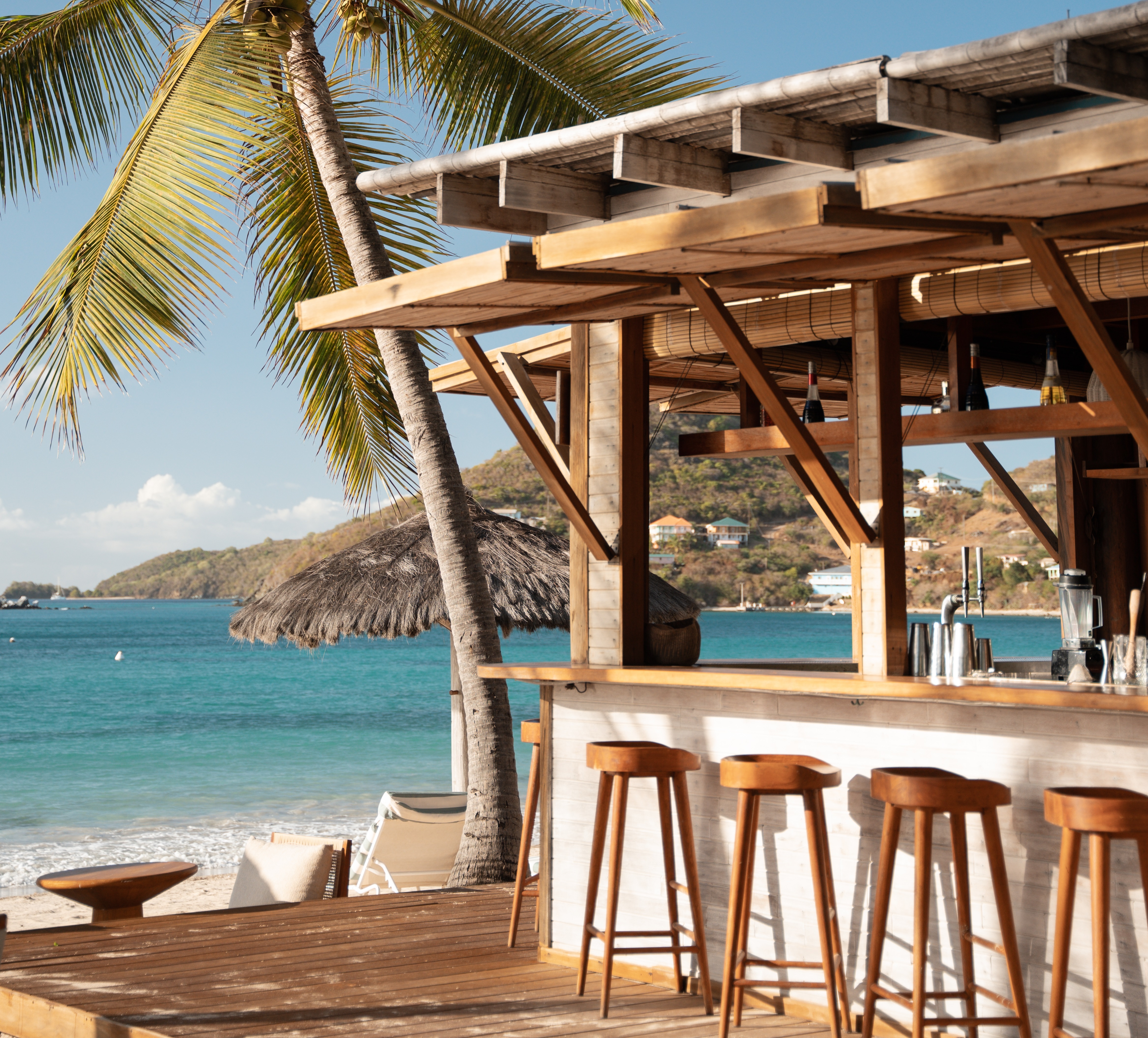 Soho Beach House Canouan view through the beach bar, with palm trees, wooden bar stools, and the sea and sand beyond