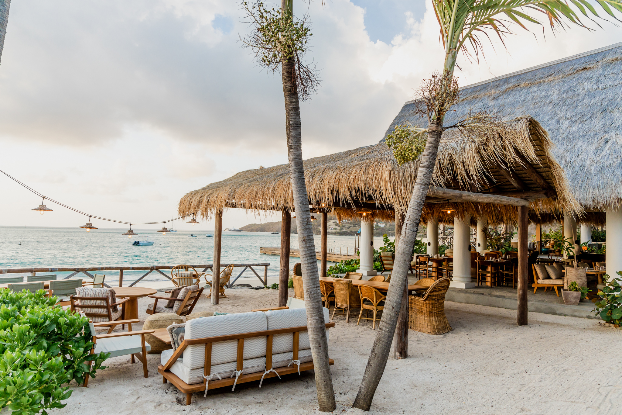 Soho Beach House Canouan beach bar with thatched roof, sofas, hanging lights, and sandy floor
