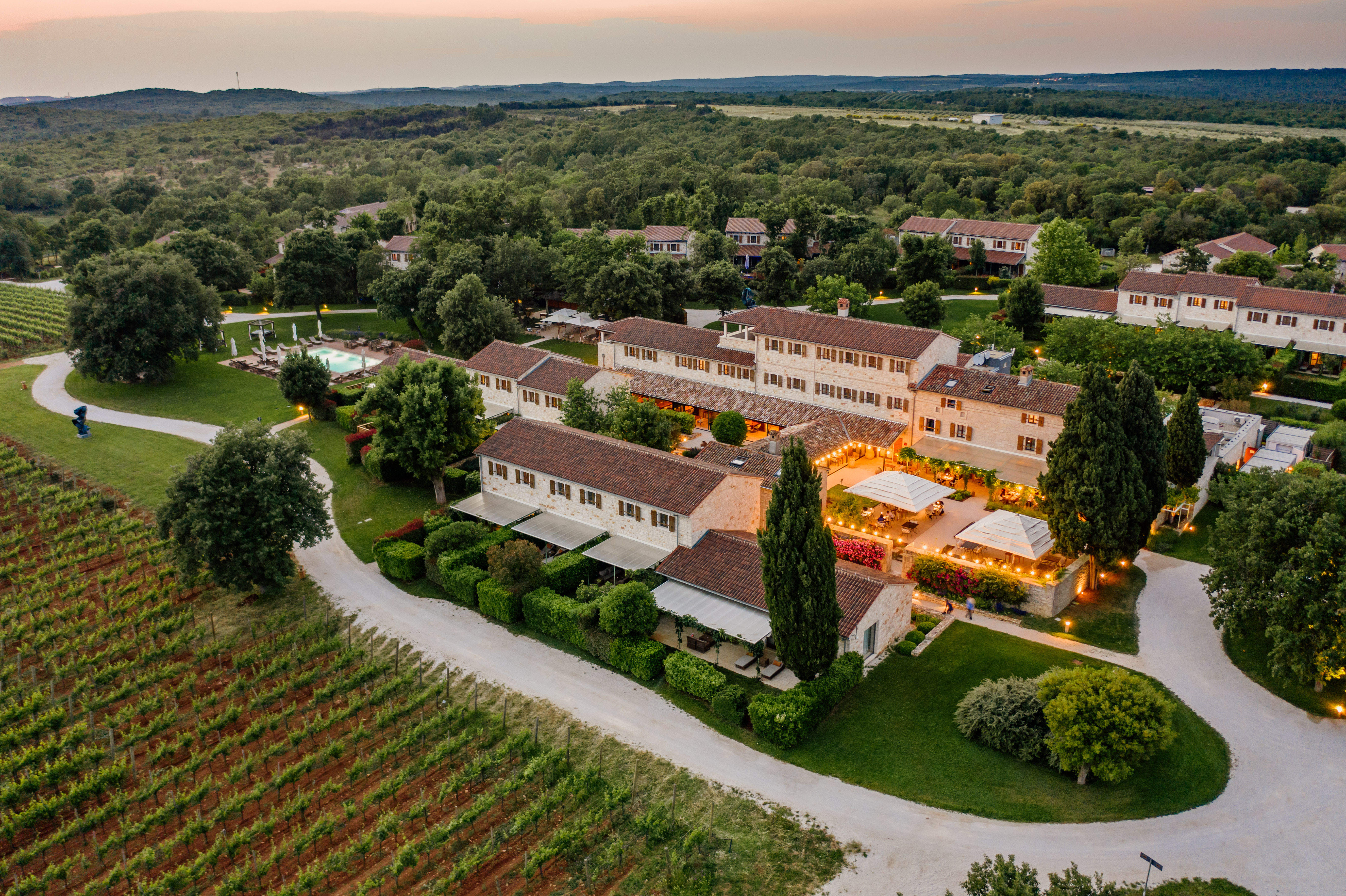 Meneghetti Wine Hotel aerial view at sunset, showing the terracotta-roofed stone buildings arranged around a central courtyard of delicately lit tables and chairs, with vineyards and forests in the distance