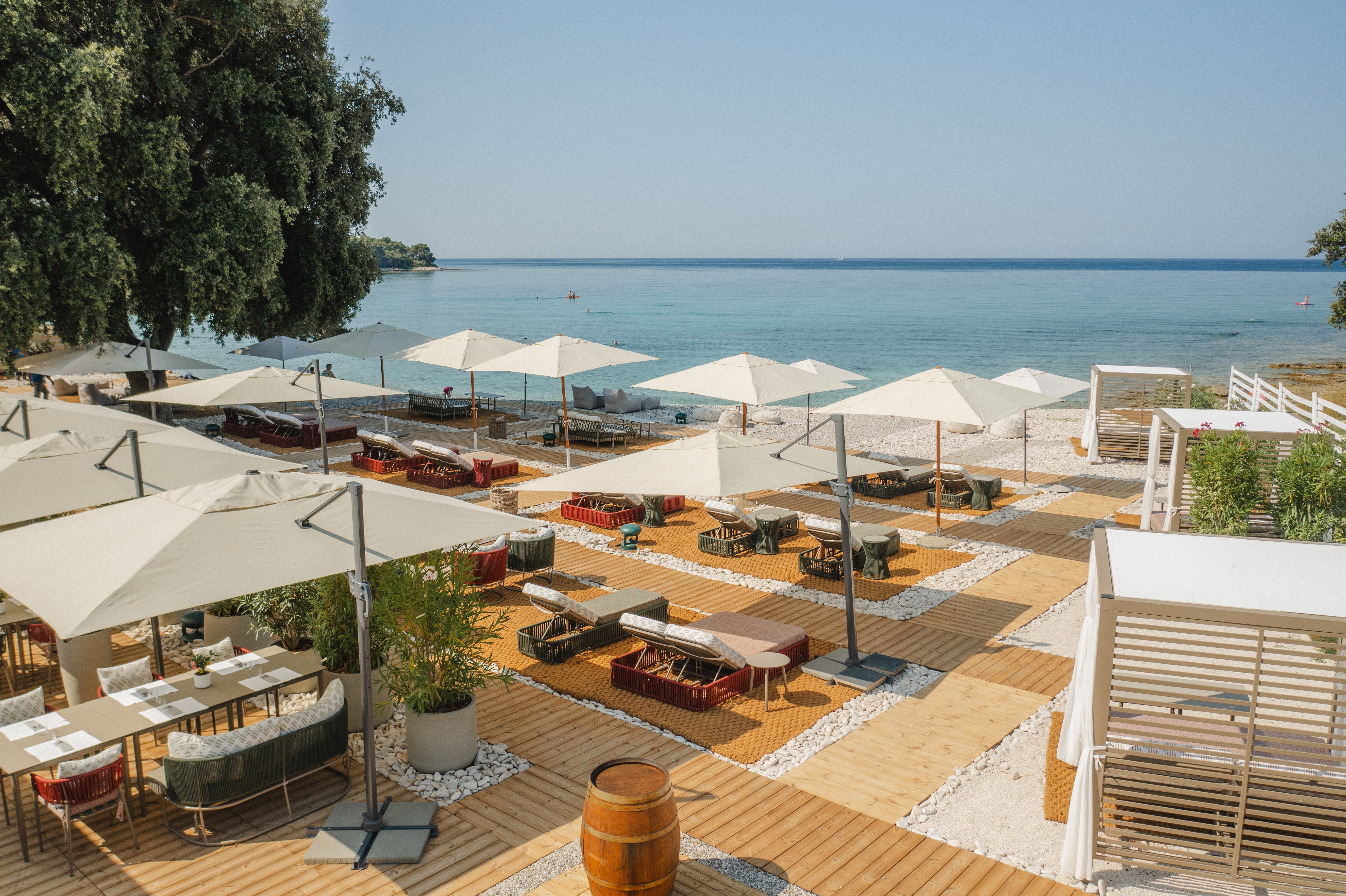 Meneghetti Wine Hotel Beach Club, with tables, chairs, and sun loungers under parasols, with wooden walkways down to the beach and blue sea beyond