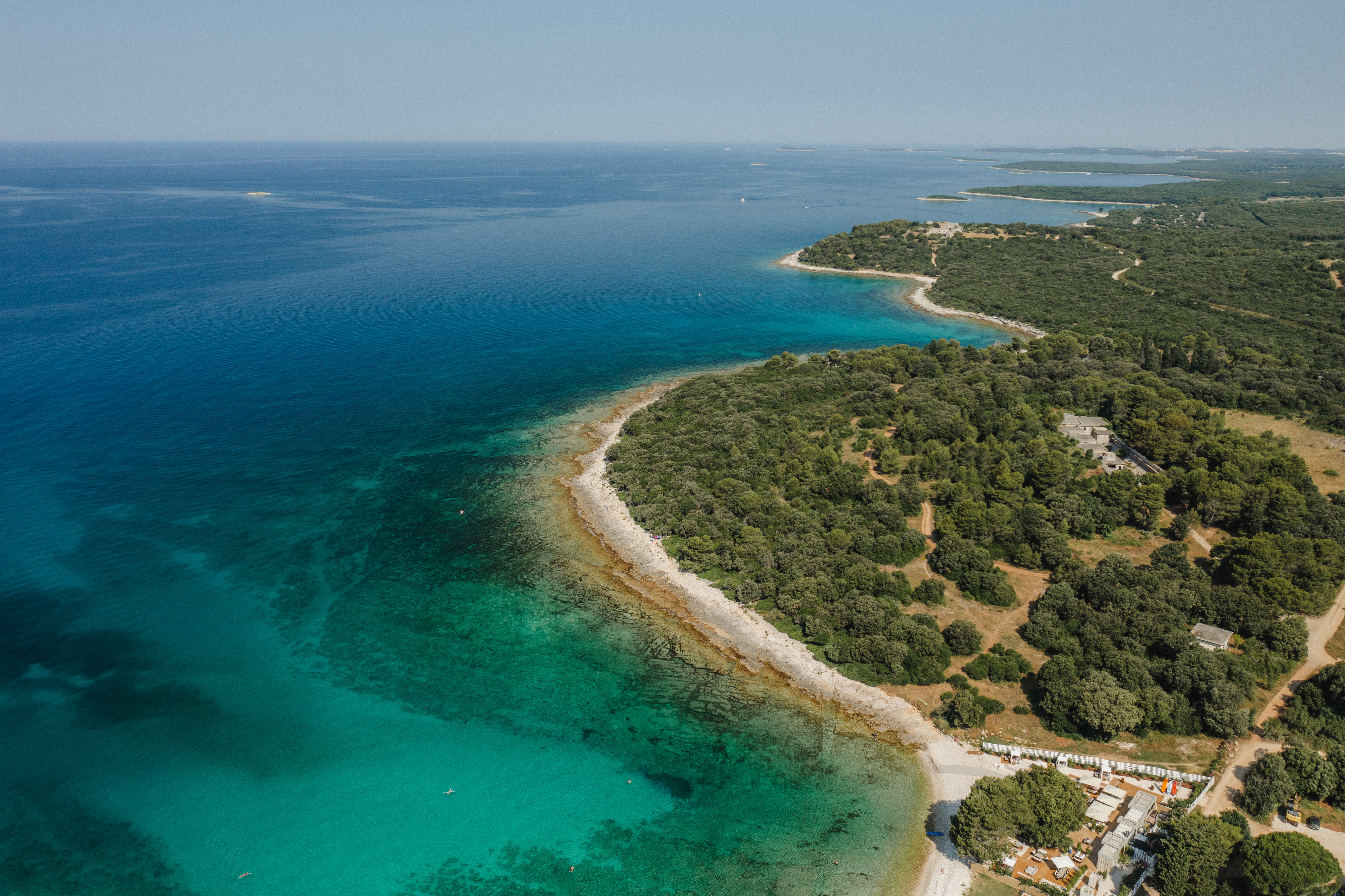 Meneghetti Wine Hotel aerial view of the Beach Club, showing bright blue sea, forested shore, and white-sandy beach