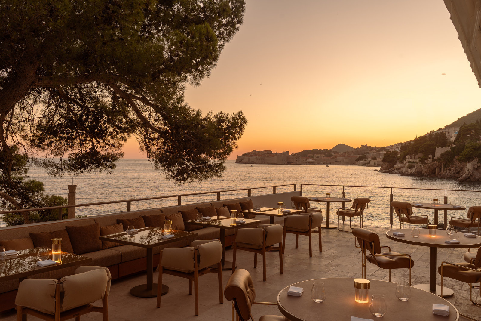 Villa Dubrovnik restaurant terrace at sunset, with tables and chairs looking out over the sea and sun disappearing behind the hills
