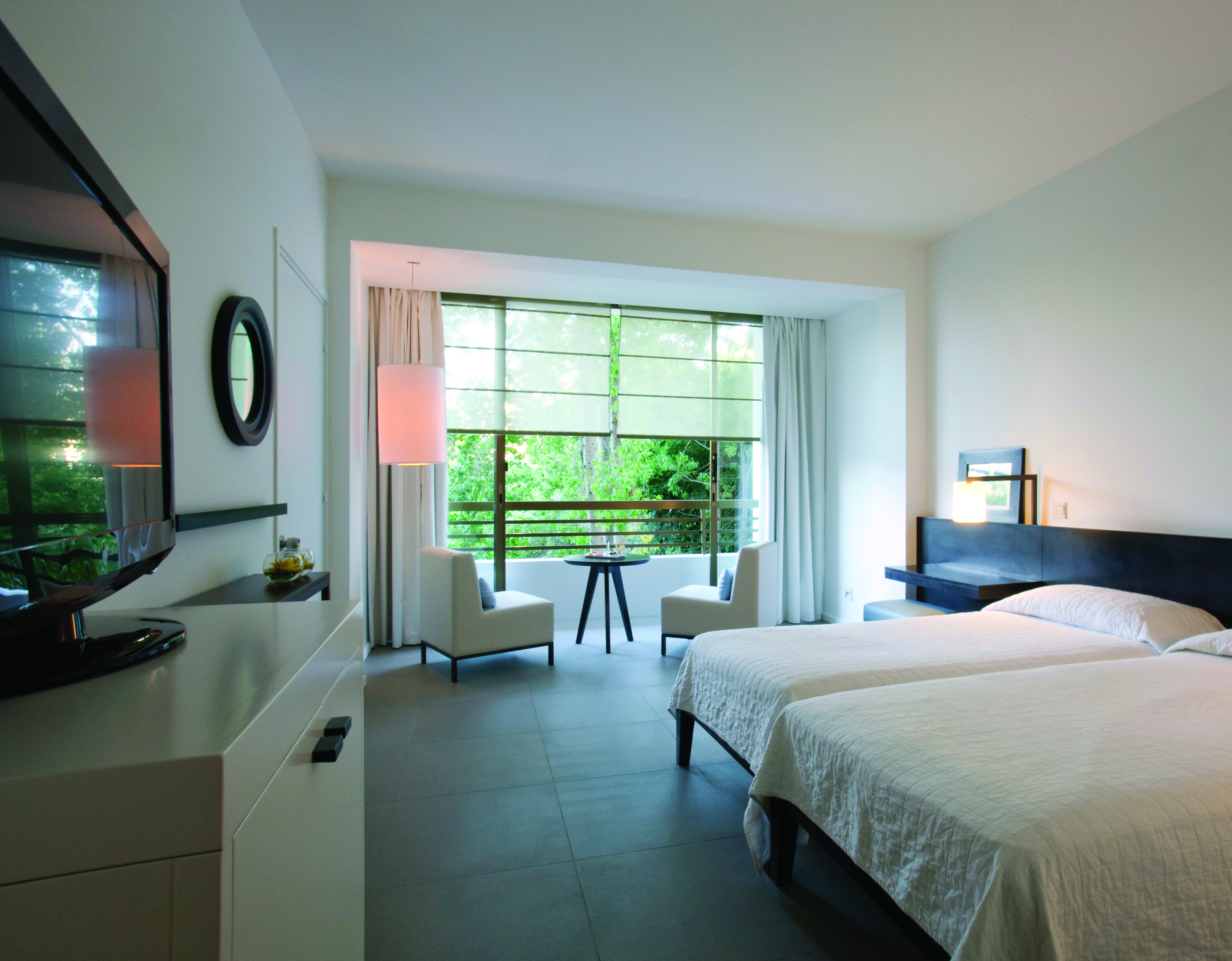 Bedroom with two beds, large glat screen television, white armchairs and large windows looking out onto greenery