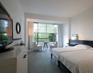 Bedroom with two beds, large glat screen television, white armchairs and large windows looking out onto greenery
