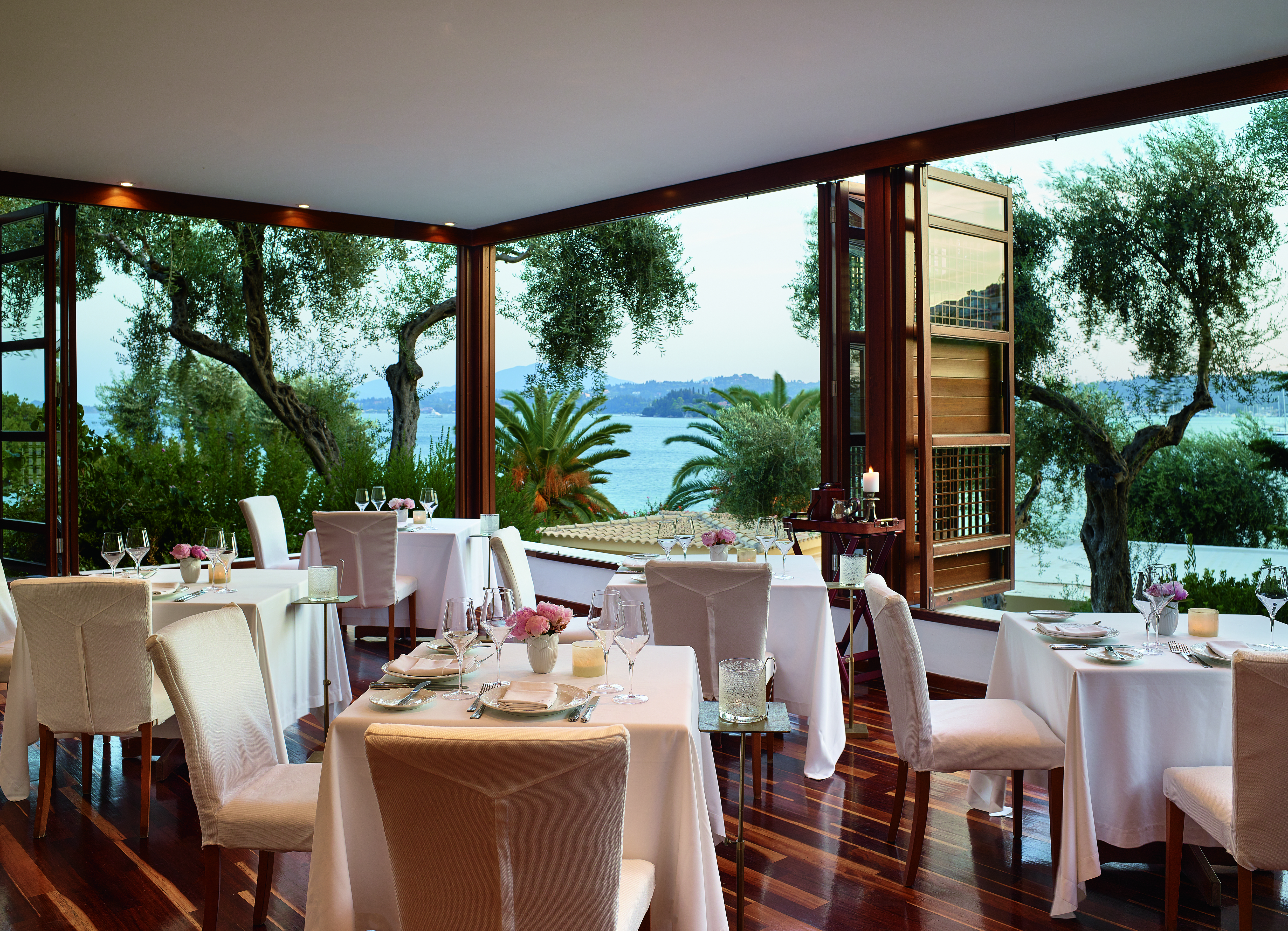 Airy restaurant with white tables and wooden floor looking onto palms
