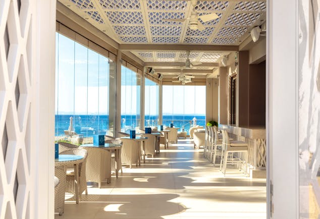 Helios bar at the Ikos Olivia with large window panneled walls looking over the ocean and white chairs and tables set up 