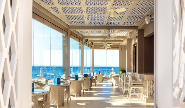 Helios bar at the Ikos Olivia with large window panneled walls looking over the ocean and white chairs and tables set up