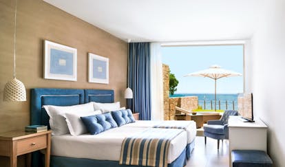 Room at the Ikos Olivia with large bed, paintings on the walls, and double doors opening up onto a terrace overlooking the sea