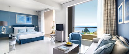 Junior suite at the Ikos Olivia with double bed, blue sofa and armchair, blue colour scheme and double doors opening up onto a seaview 