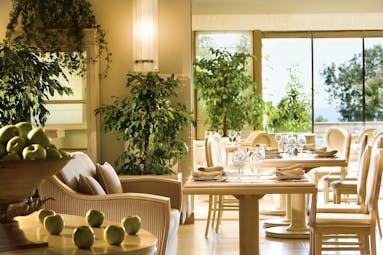 View of the Veranda restaurant at the Sani Beach. Dining chairs and tables are set up around the room with hanging plants and potted trees 