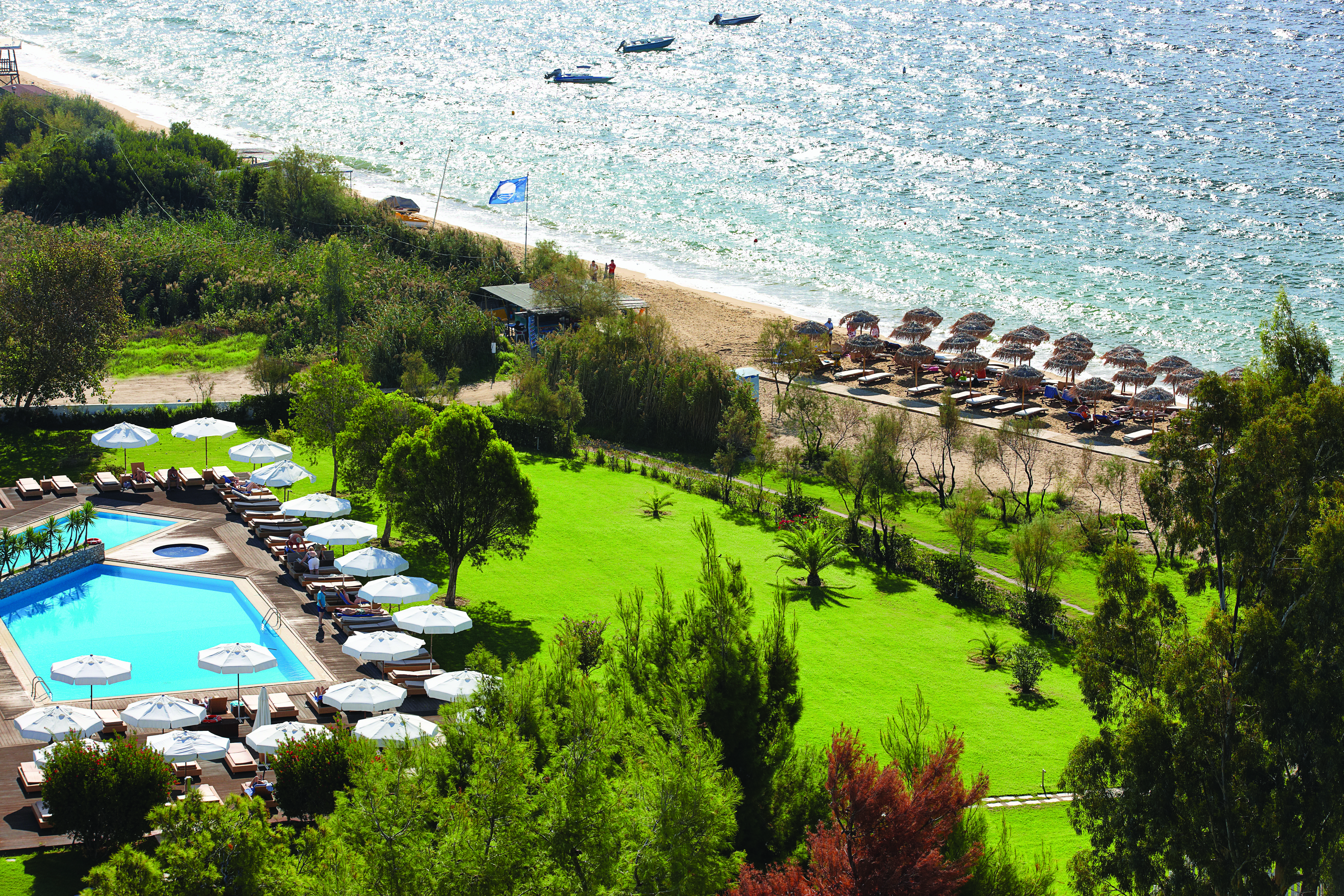 Overview of the Skiathos Princess hotel showing a large pool surrounded by white umbrellas and deck chairs. The beach is shown in close proximity