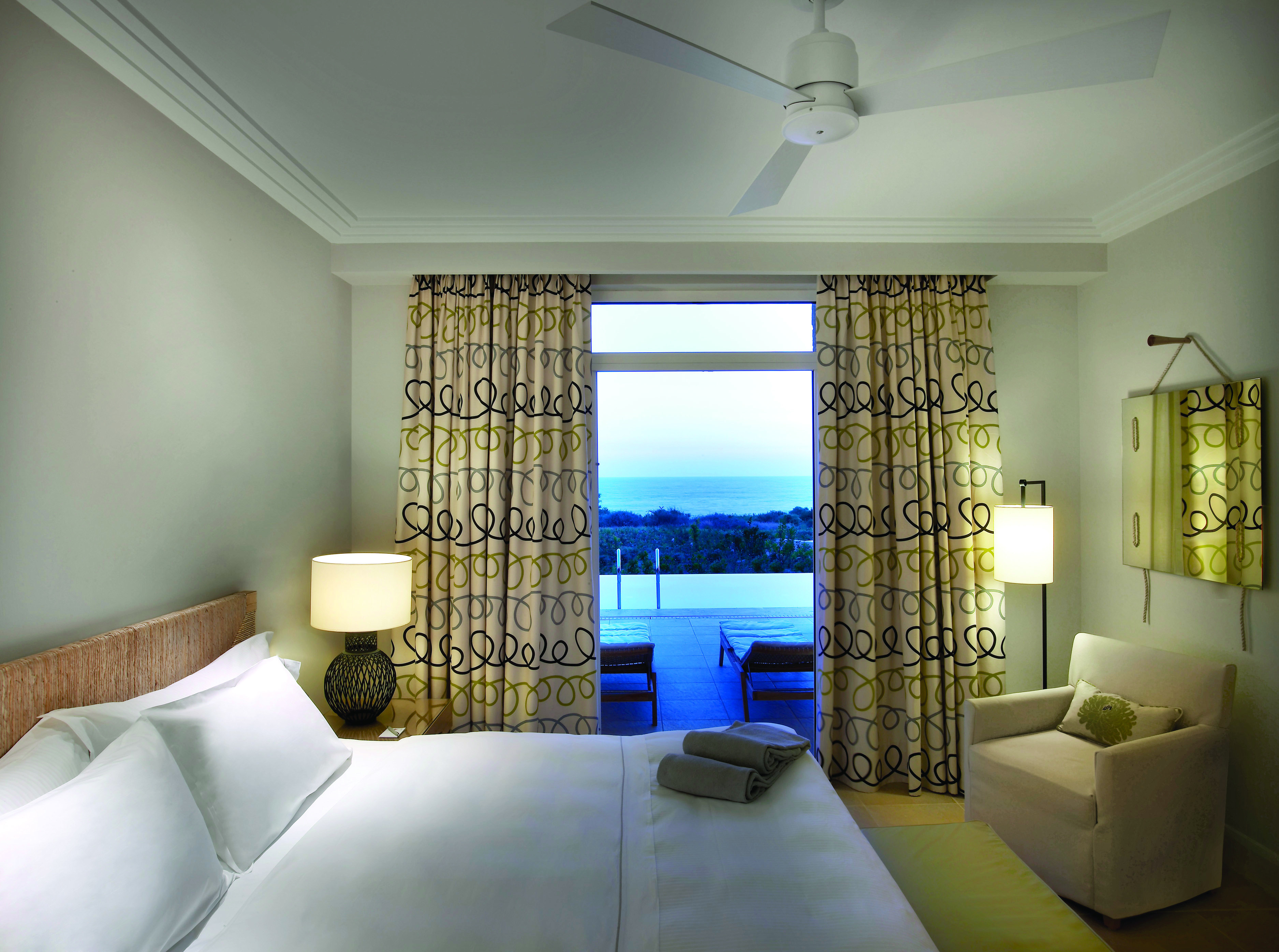Bedroom at the Westin Resort with big double bed, armchair, and window opening onto a terrace with an infinity pool