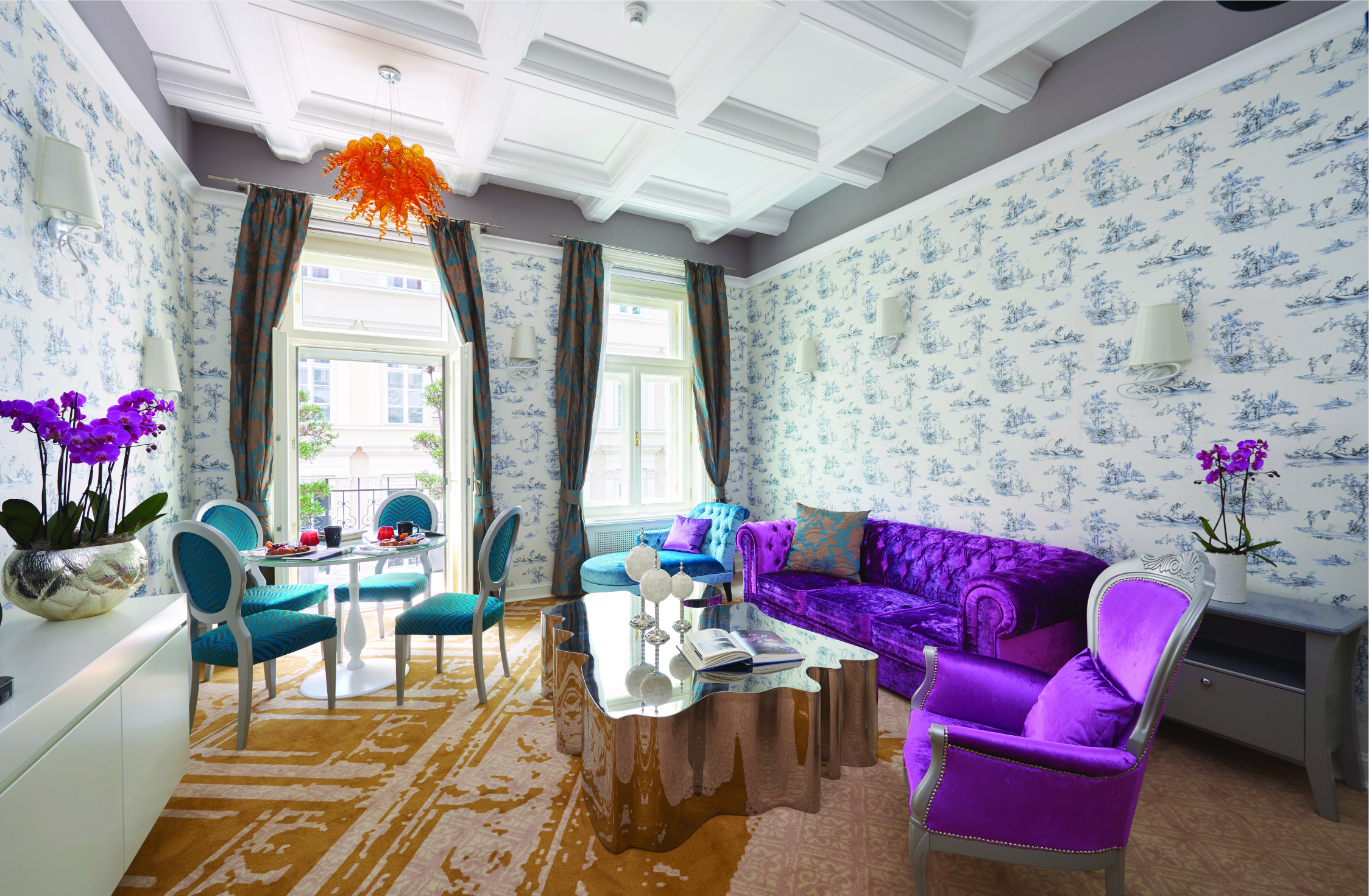 Aria Hotel Budapest suite sitting room  orange chandelier purple sofa and chair blue chaise longue table and blue chairs 