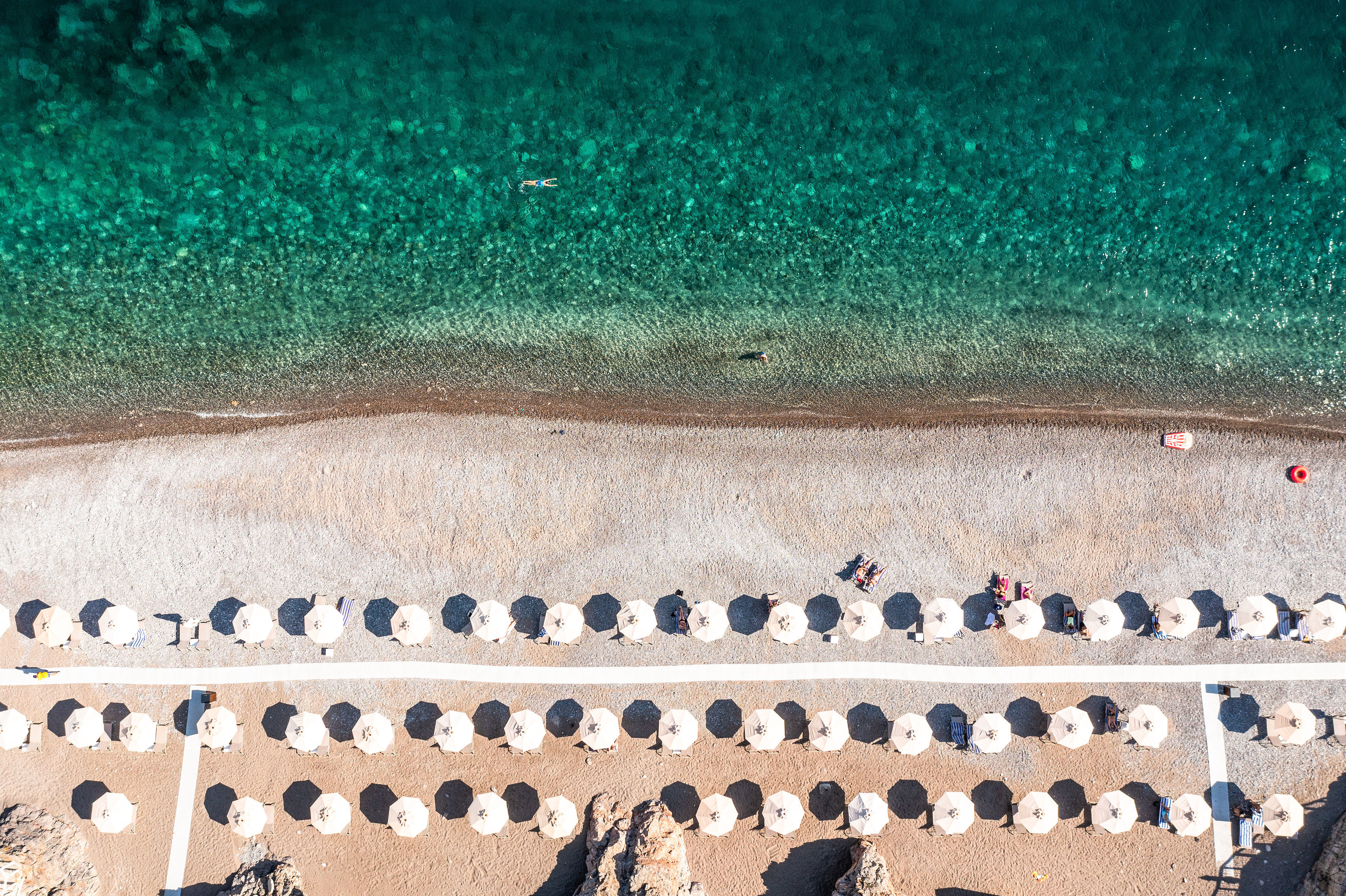 Chedi Lustica Bay beach seen from above, with crystal clear blue waters and white sand dotted with parasols