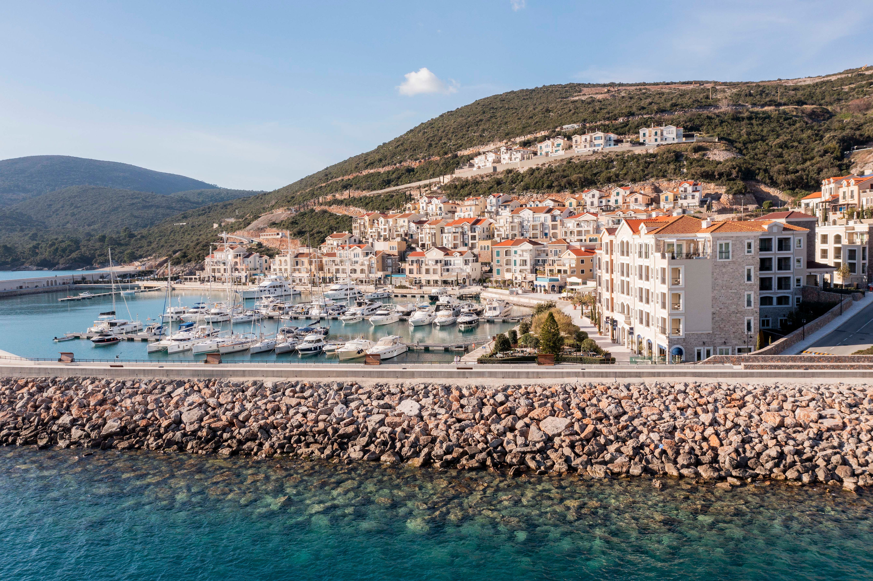 Chedi Lustica Bay marina, with promenade and rows of sailing boats and yachts, and the terracotta roofs of the surrounding town visible climbing up the hillside
