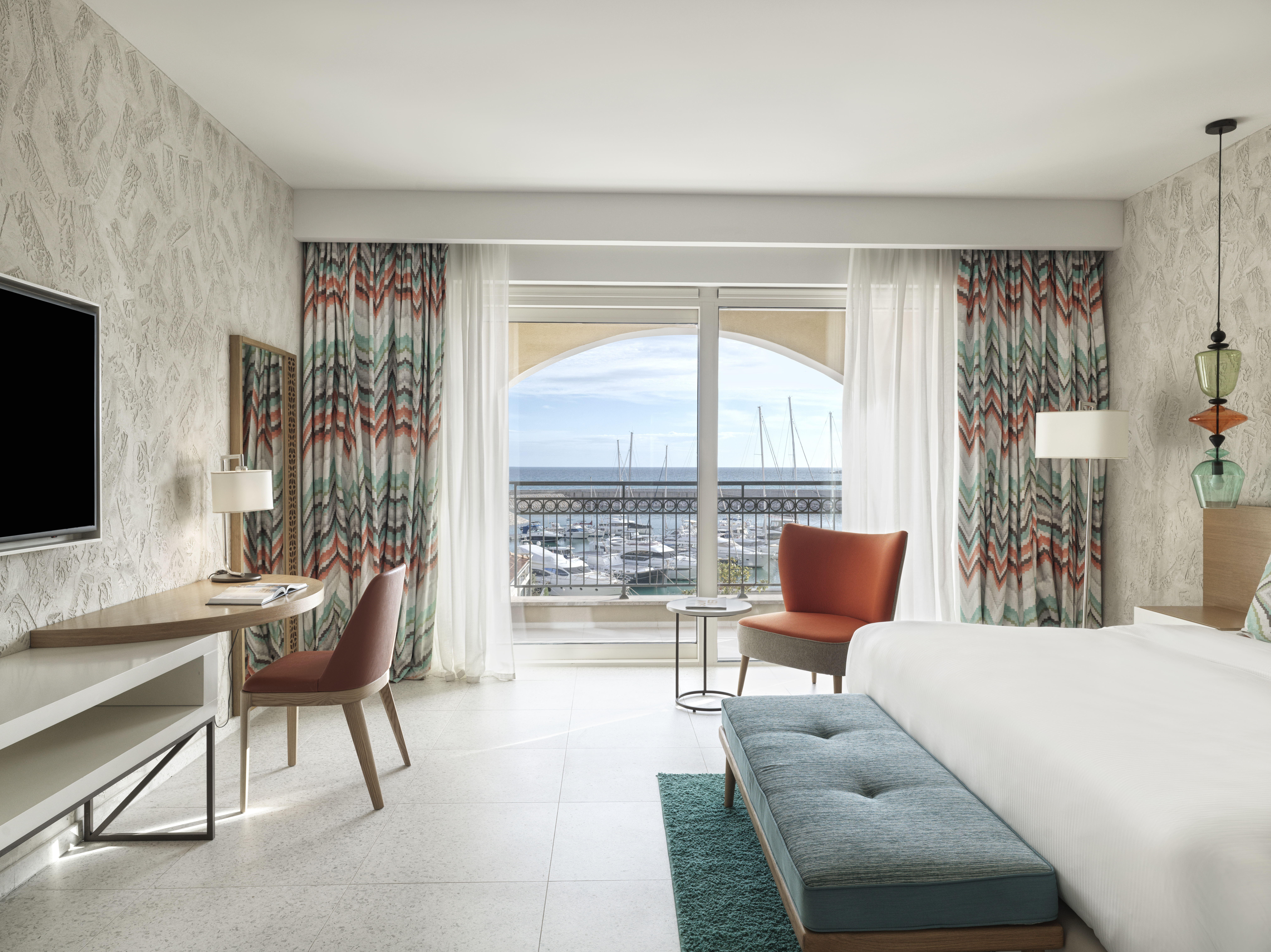Chedi Lustica Bay room with light patterned wallpaper, white linens, and colourful touches in the curtains and soft furnishings, and bright views of the marina