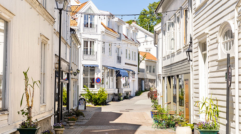 Home Hotel Grimstad location, showing a narrow pedestrianised street with white wooden houses on either side, plants outside the front doors, and orange tiled roofs visible