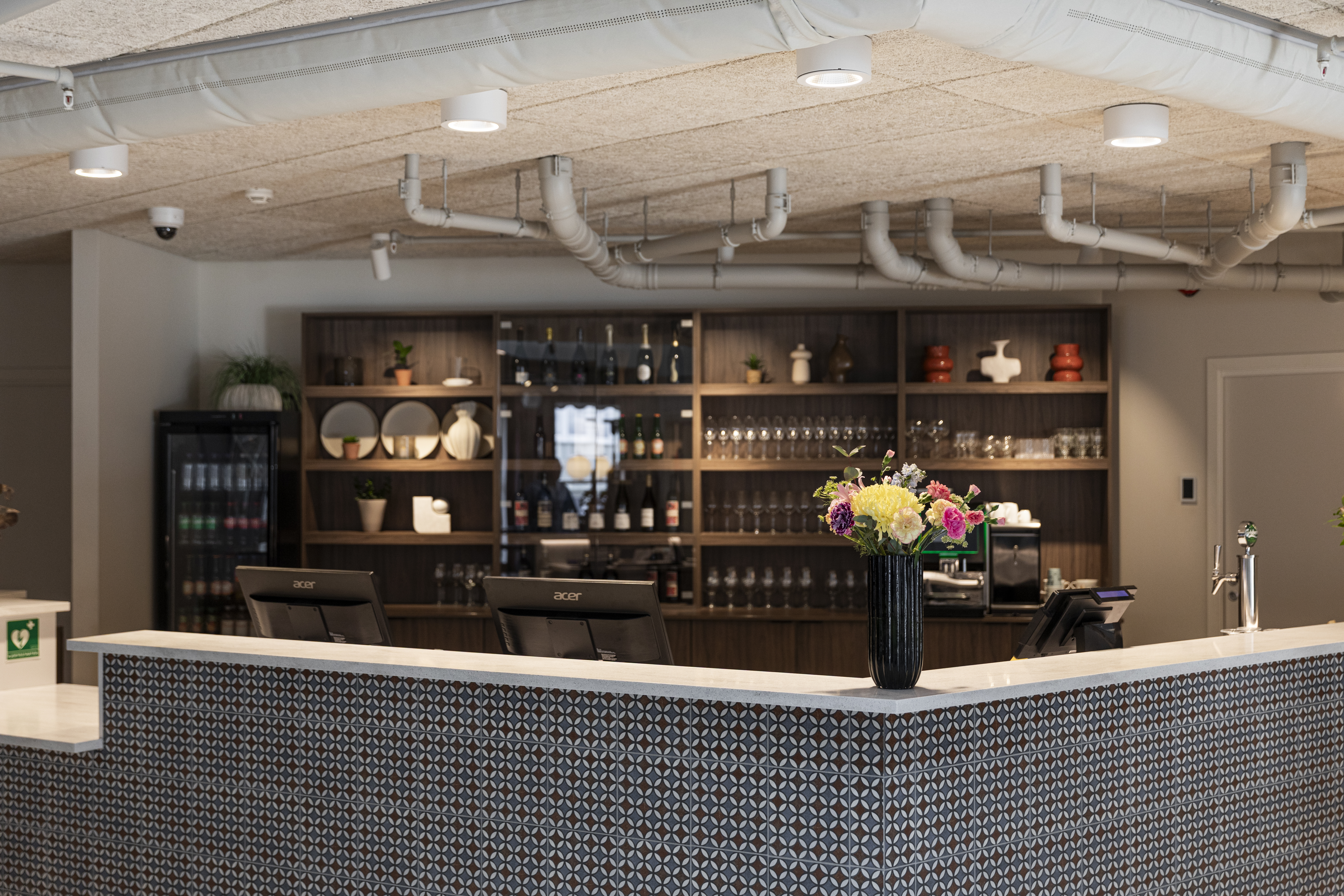 Home Hotel Grimstad Reception, with tiled black and white desk, flowers, and wooden shelves of bottles, plates, and glasses on the back wall