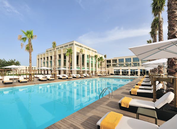 View of the exterior of the hotel with the swimming pool infront, palm trees and white sun loungers