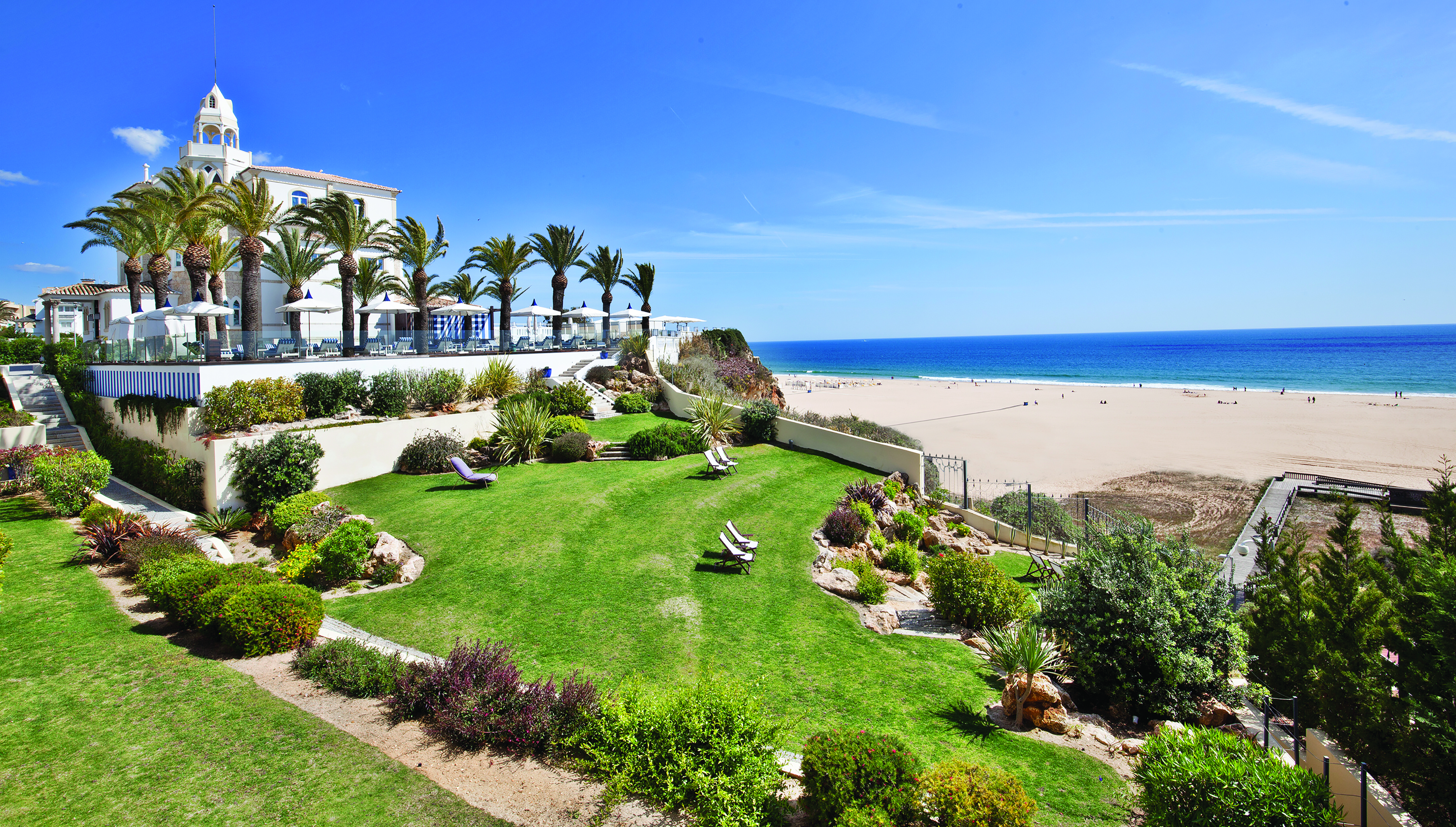 View of the hotel overlooking a sandy beach with palm trees and sunbeds surrounding it