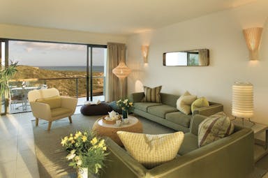 View of a bay house standard room looking over rocks and the sea with big grey sofas and flowering decorating the room