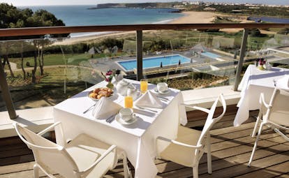 view of the terrace used for serving breakfast, with tables and chairs set up on a terrace balcony looking over a blue pool and sandy beach