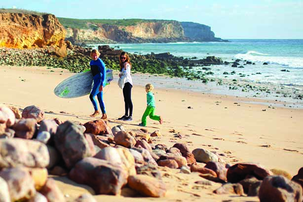 Shows a family on the beach near water holding a surf board 