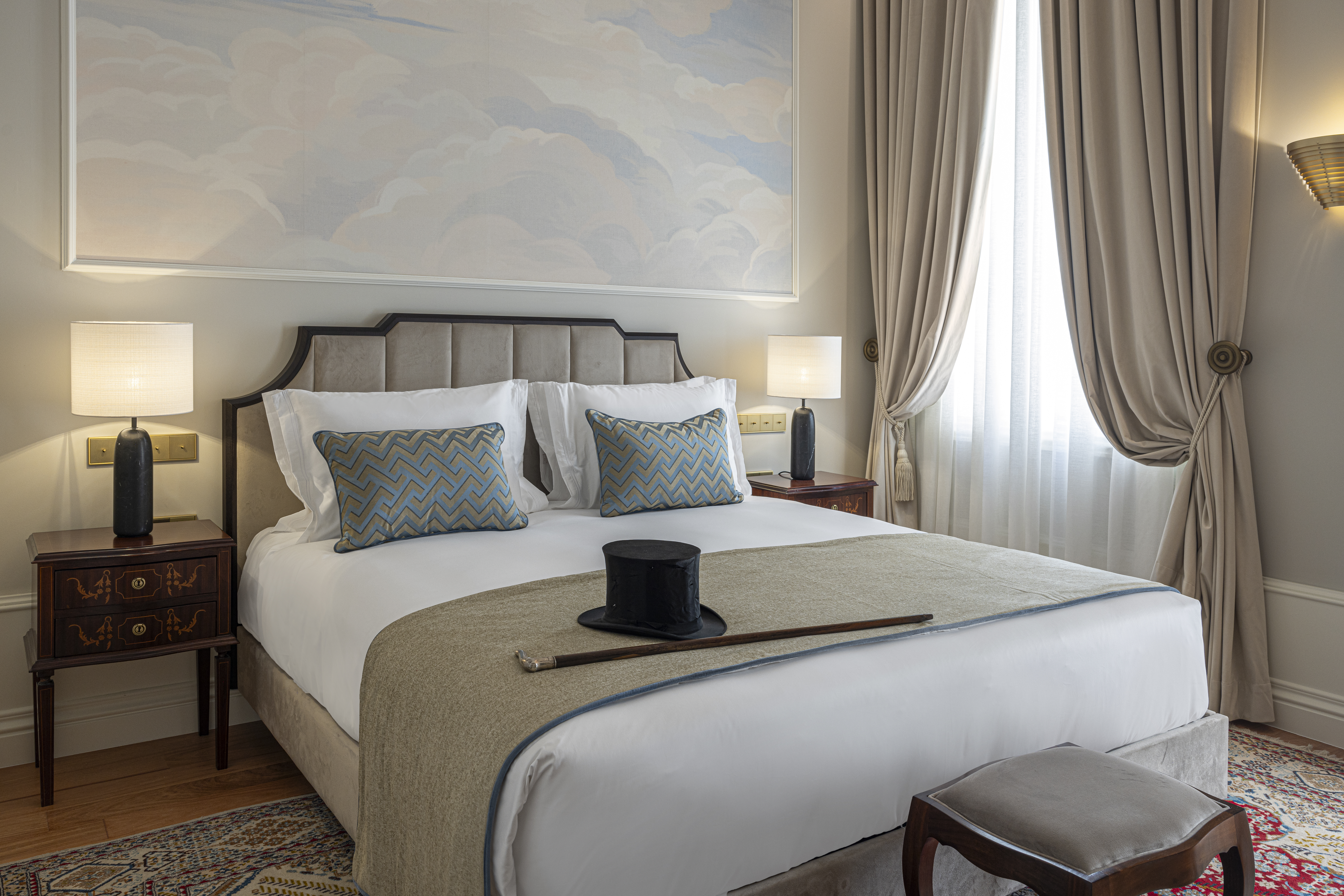 Palacete de Valdemouro Artur Corvelo themed room, with neutral colour palette, draped curtians, plush headboard, soft mural on the wall, and top hat and cane on the bedspread