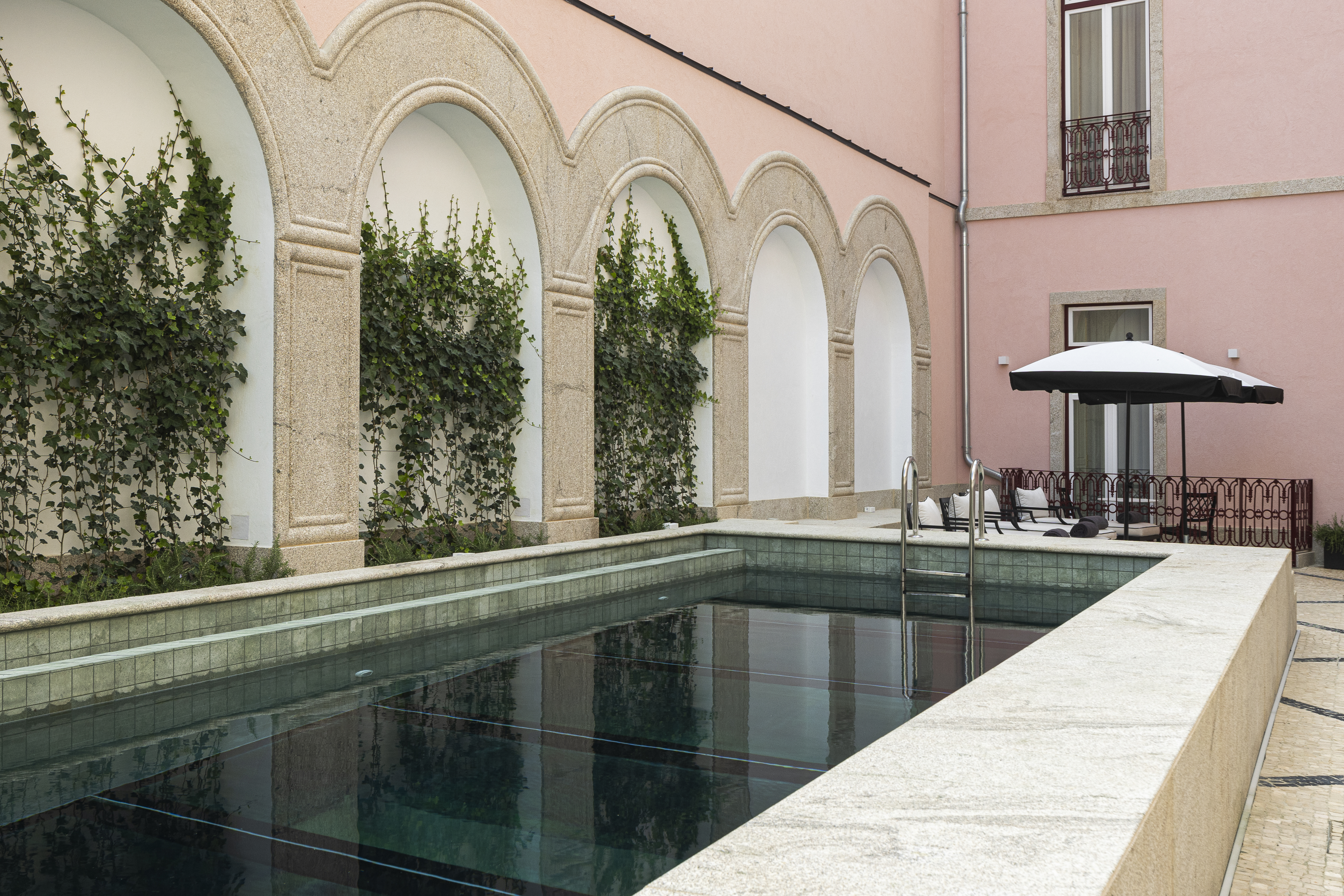 Palacete de Valdemouro pool, with marble surround, sun loungers and parasols on the far side, pink stone walls, and arches cutting into the walls housing climbing vines
