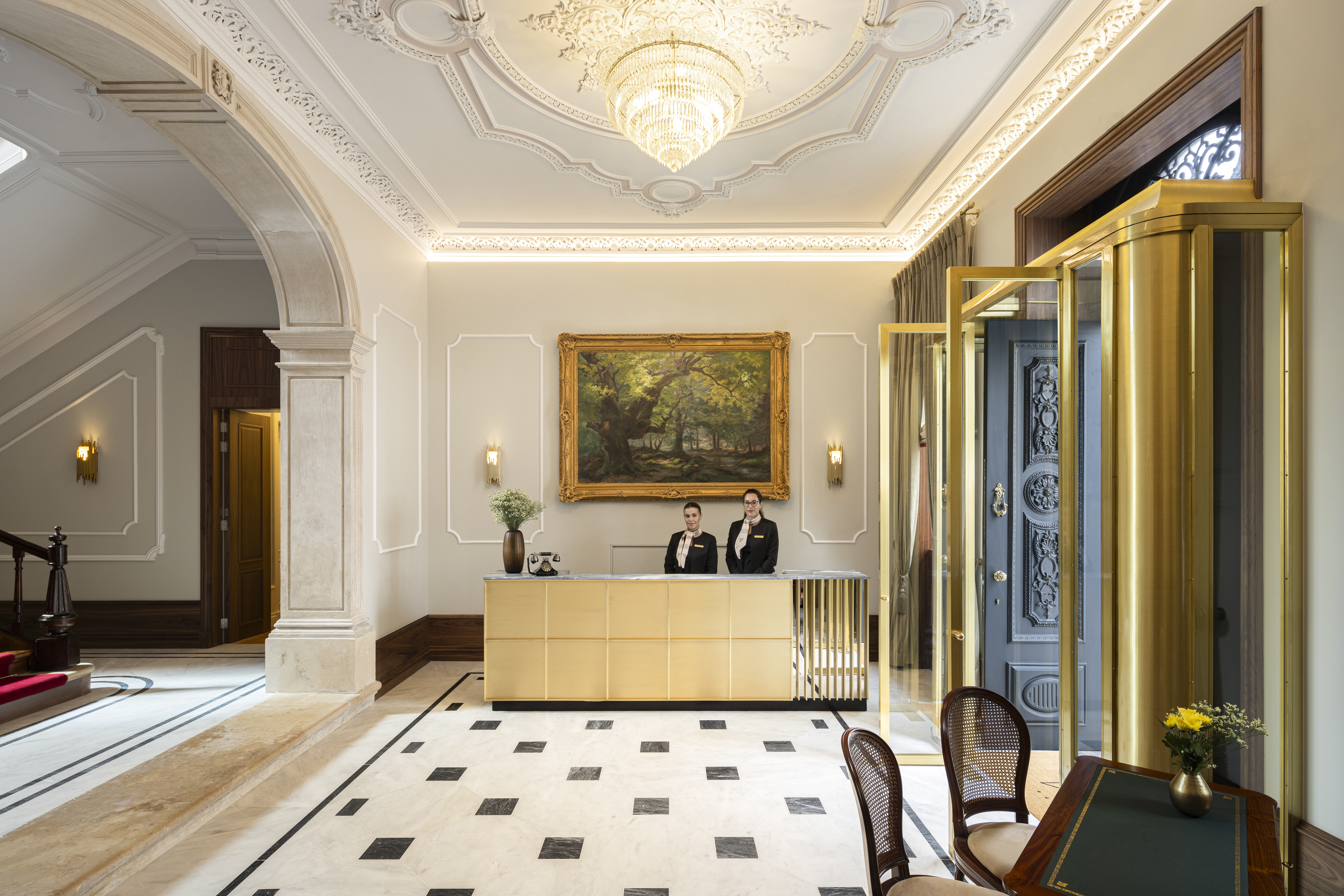 Palacete de Valdemouro reception, with two figures stood behind the light-wood desk, gold-framed artwork behind them, gold front doors, an archway leading towards a red-carpeted staircase, and an ornate ceiling design