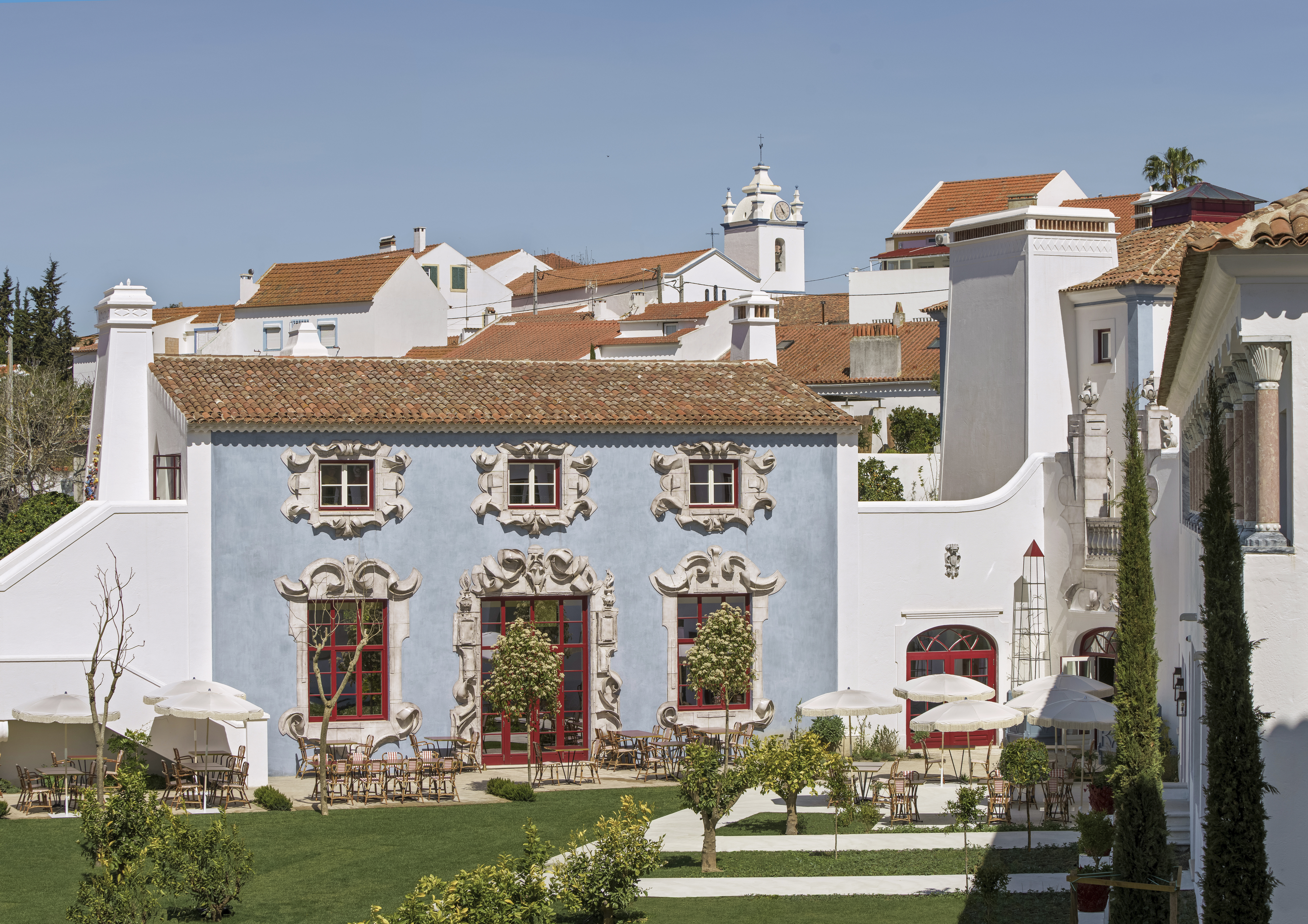 Vermelho Melides exterior, with blue walls and ornate white and red window frames, gardens in the foreground, and terracotta rooves stretching beyond