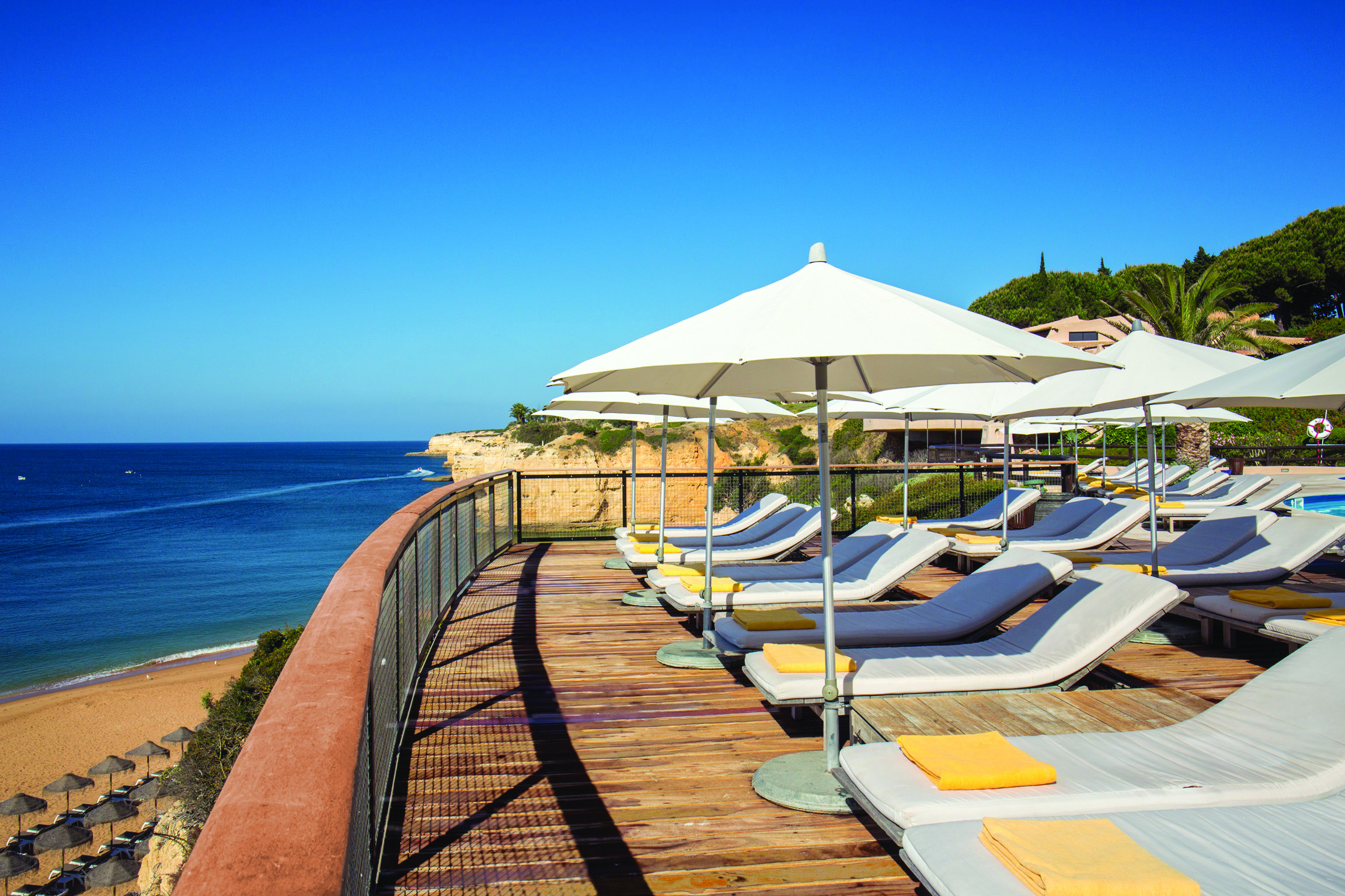 View of the pool deck looking over the beach with white umbrellas and sunbeds on a wooden deck 