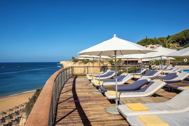View of the pool deck looking over the beach with white umbrellas and sunbeds on a wooden deck 