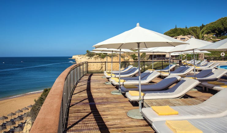 View of the pool deck looking over the beach with white umbrellas and sunbeds on a wooden deck 