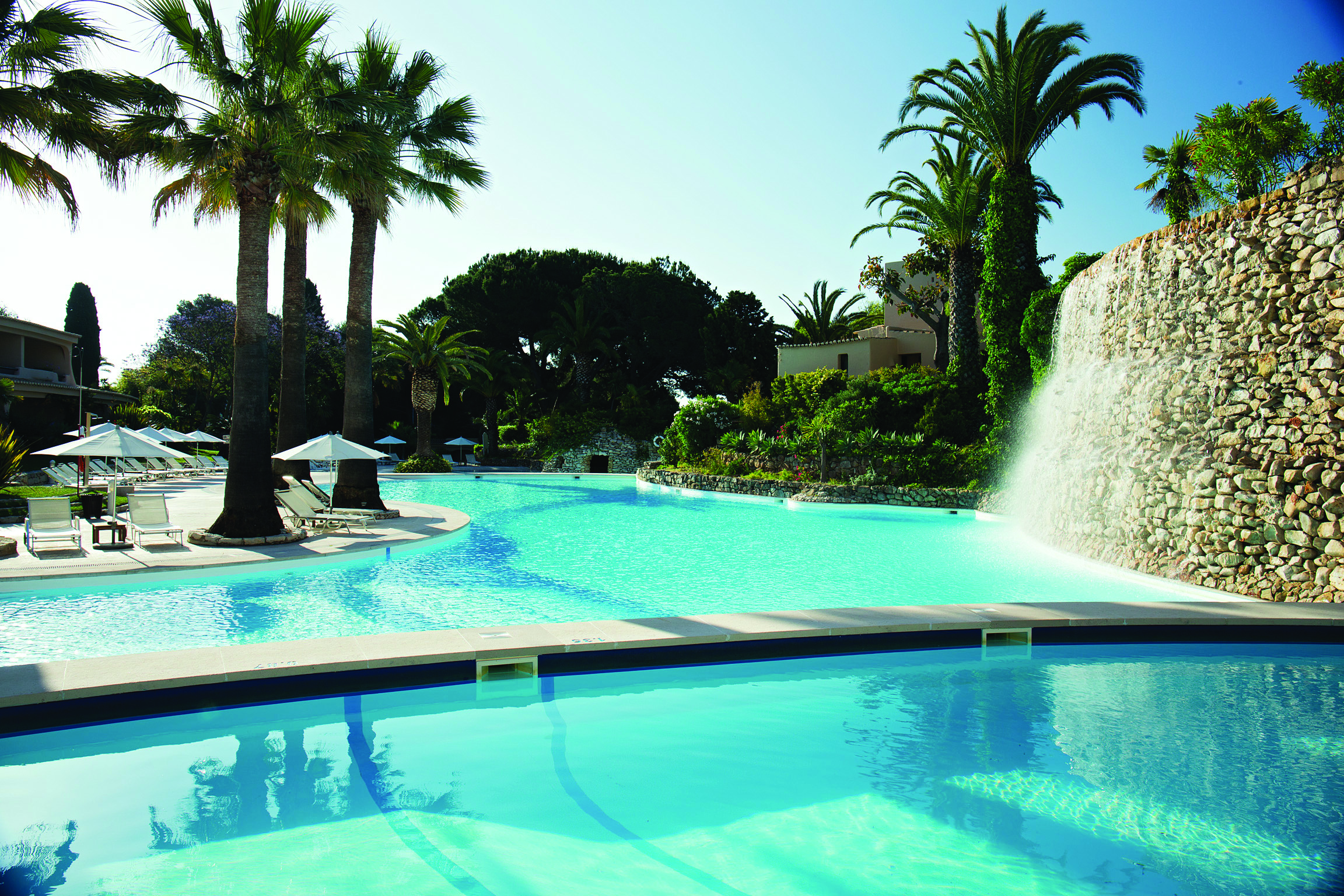 View of the swimming pool at the vilalara thalassa resort with palm trees and white umbrellas around the edge
