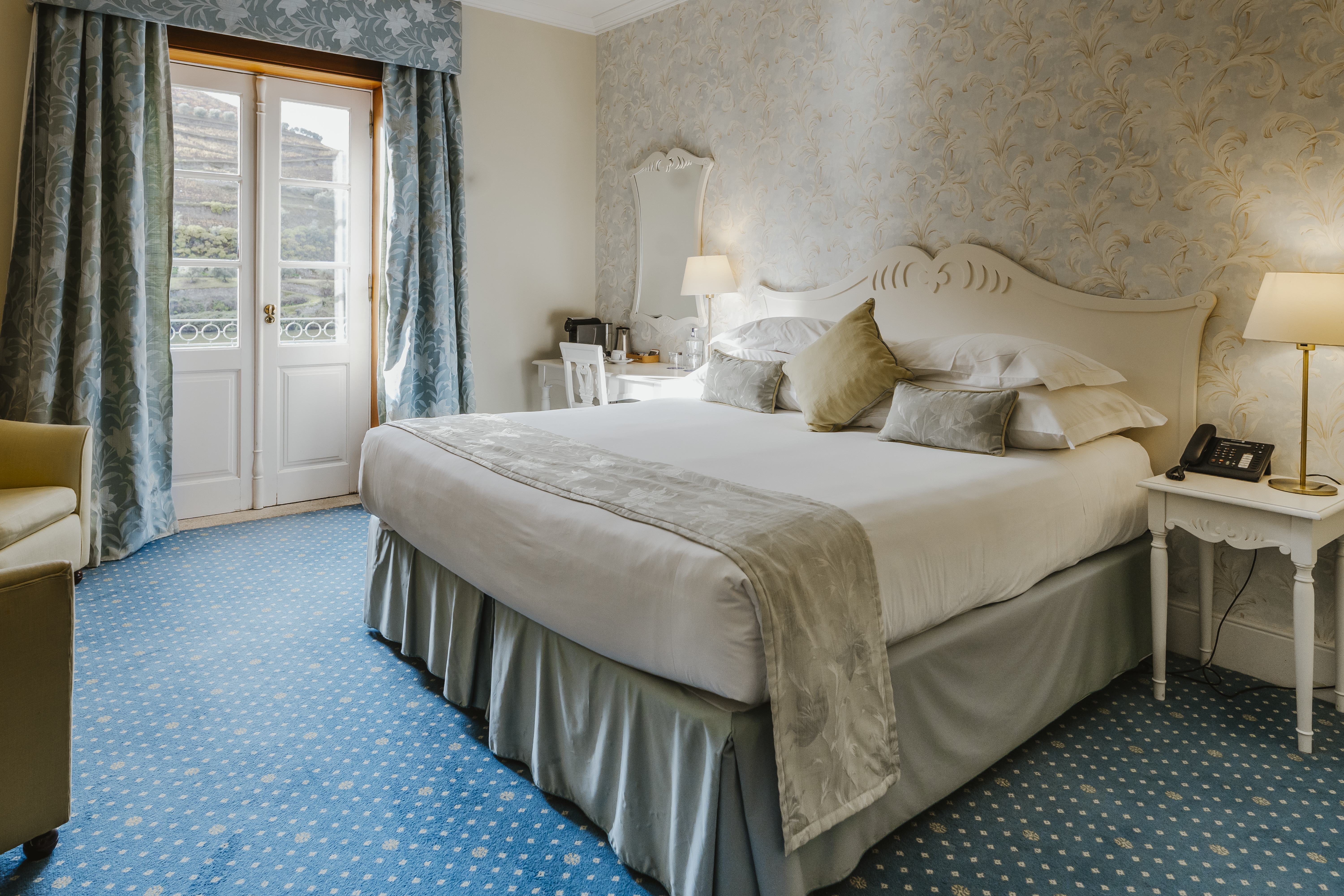 Vintage House Pinhao Standard Room with grand, ornate furnishings, patterned neutral wallpaper, and double doors opening up onto river and hill views